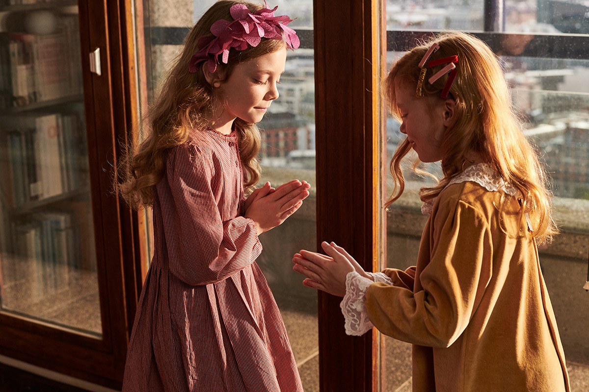 Two young girls wearing dresses stand near a window. One is in a pink dress with a floral headband; the other is in gold.