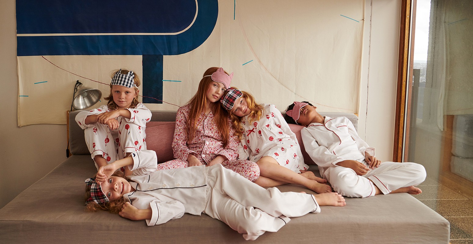 Group of five children in matching patterned pyjamas lounging on bed with sleep masks, bathed in natural light in an indoor setting.