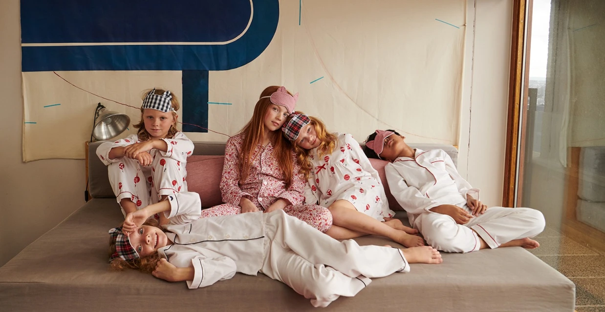 Group of five children in matching patterned pyjamas lounging on bed with sleep masks, bathed in natural light in an indoor setting.