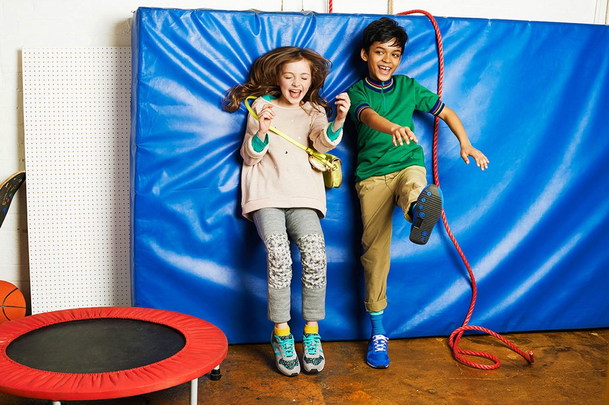Two smiling kids, a boy and a girl, are playing against a blue padded wall with a red trampoline visible nearby in an indoor setting.