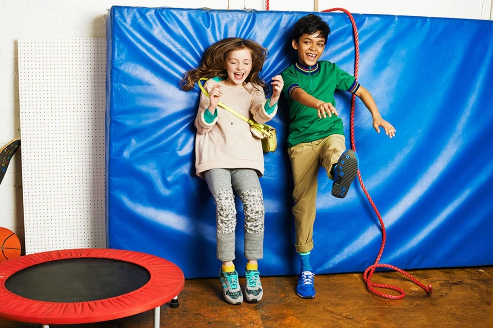 Two smiling kids, a boy and a girl, are playing against a blue padded wall with a red trampoline visible nearby in an indoor setting.