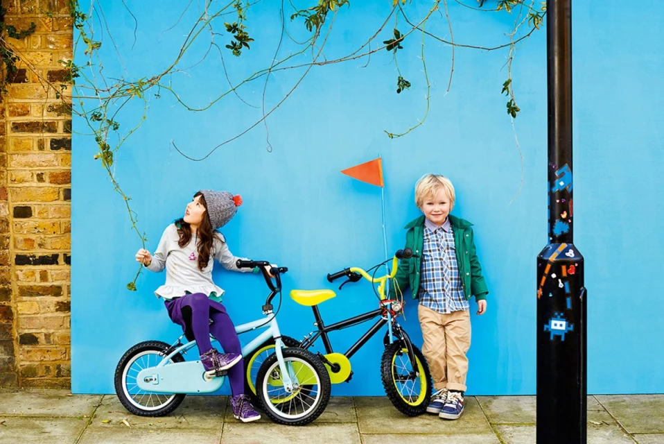 Two children, a boy and a girl, pose with their bicycles against a bright blue wall with climbing vines, creating a colorful, playful scene.