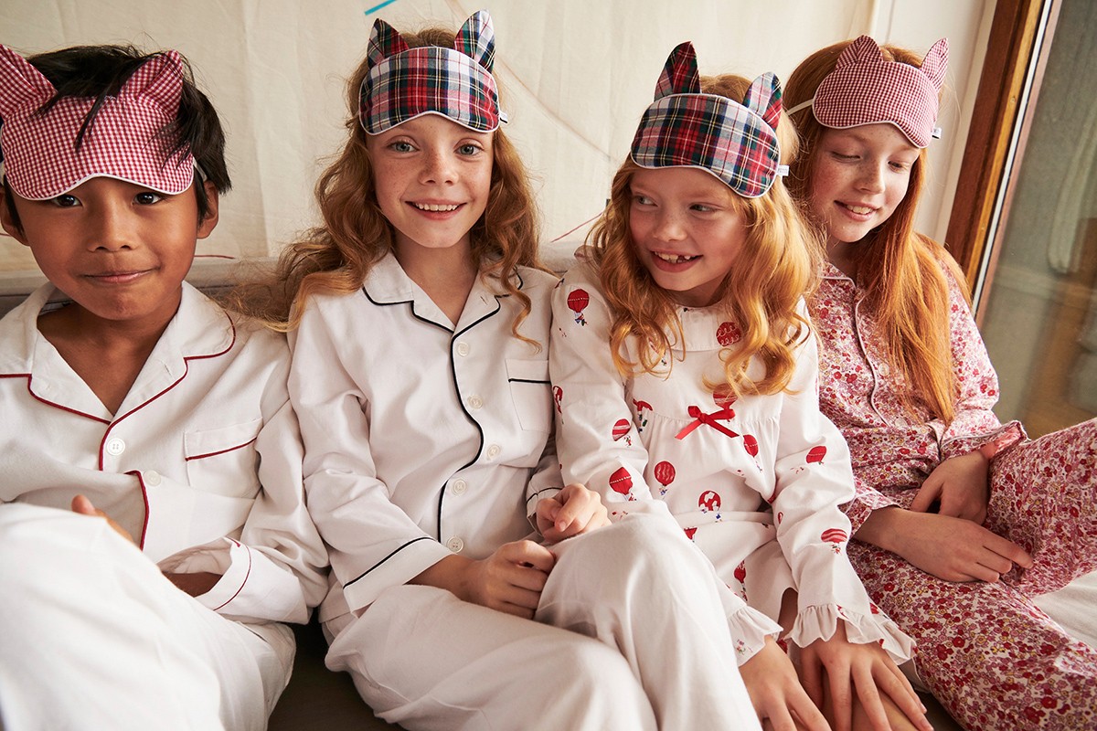 Four children in matching pajamas and sleep masks sit close together indoors, smiling at the camera in a brightly lit room.