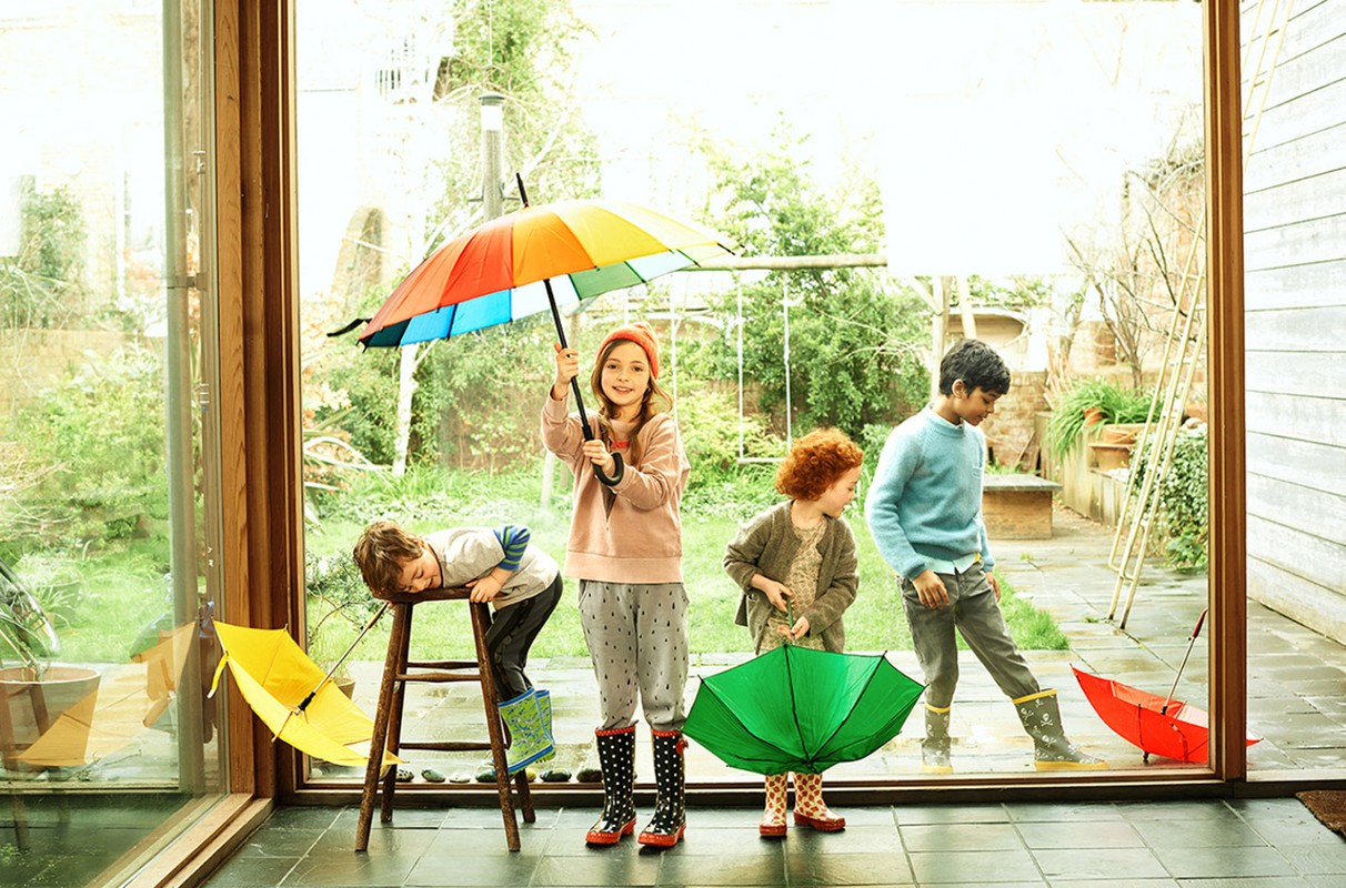 Four children playing inside near a window with a colorful and green umbrellas, wearing rain boots and casual clothing on a rainy day.