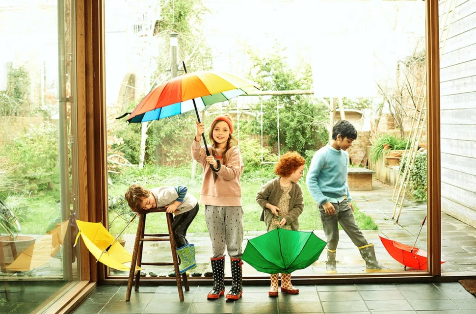 Four children playing inside near a window with a colorful and green umbrellas, wearing rain boots and casual clothing on a rainy day.