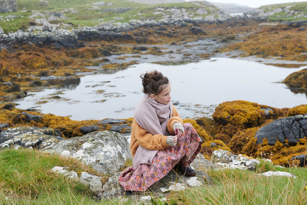 Young girl wearing an oversized orange knit sweater and scarf crouching on rocky coastal terrain by water