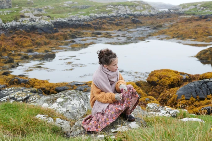 Young girl wearing an oversized orange knit sweater and scarf crouching on rocky coastal terrain by water