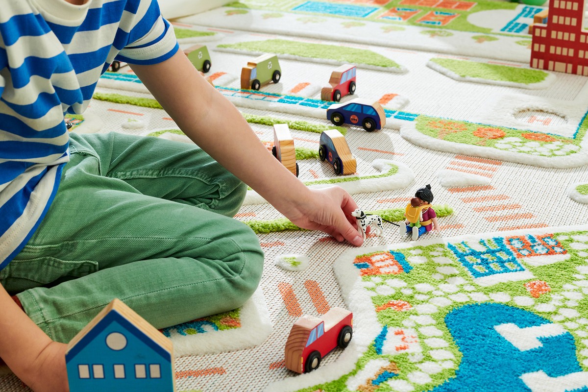 Child plays with toy cars and figures on a city-themed rug indoors, showcasing a playful and imaginative scene.