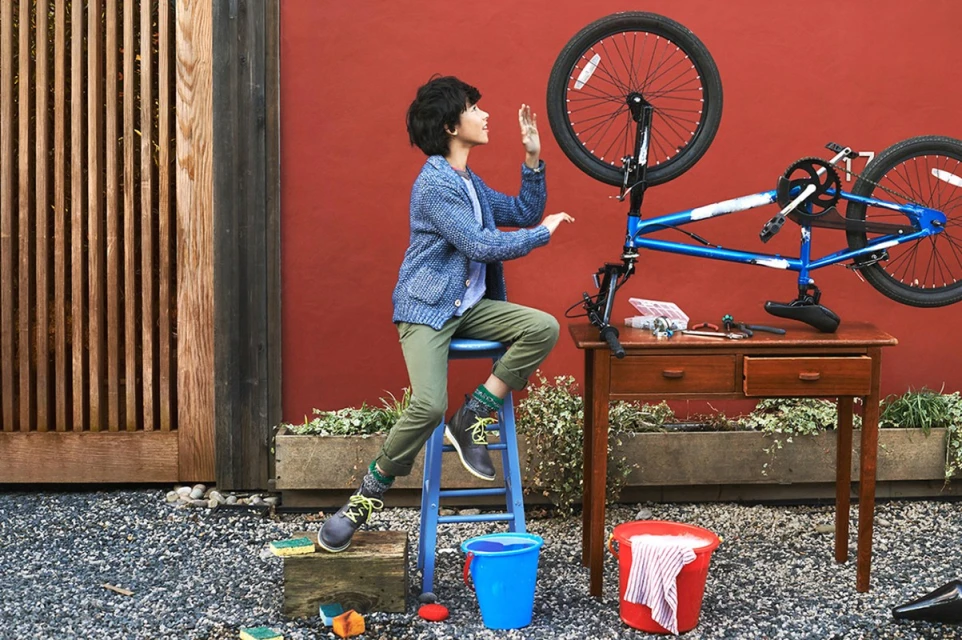 Young person repairs a bicycle outside on a gravel surface near a wooden fence and brick wall, using a small wooden table.