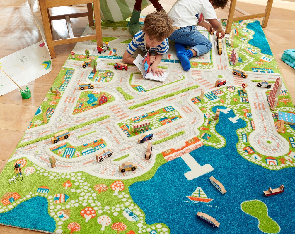Two young children play on a colourful rug with a road and town pattern, surrounded by toy cars and wooden blocks in an indoor setting.