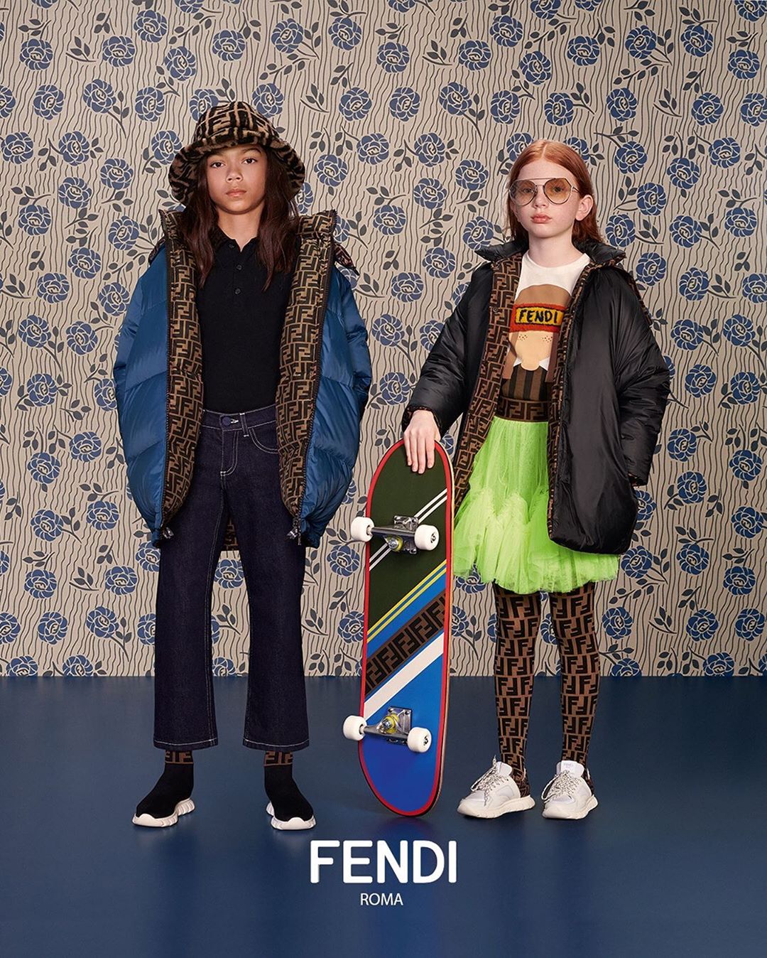 Two children pose in Fendi apparel; one holds a skateboard, set against a floral backdrop in a fashion editorial style image.