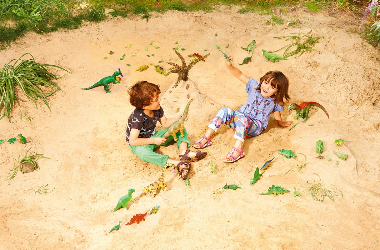 Two children play with toy dinosaurs in a large sand pit, arranged around a sandcastle volcano surrounded by lush green plants.