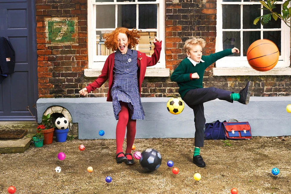 Two children, a red-haired girl and a blonde boy, playing with an assortment of balls in a yard with a brick house behind them.
