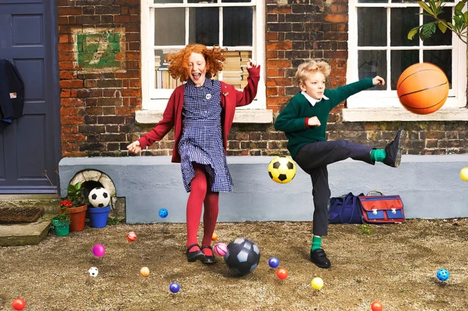 Two children, a red-haired girl and a blonde boy, playing with an assortment of balls in a yard with a brick house behind them.