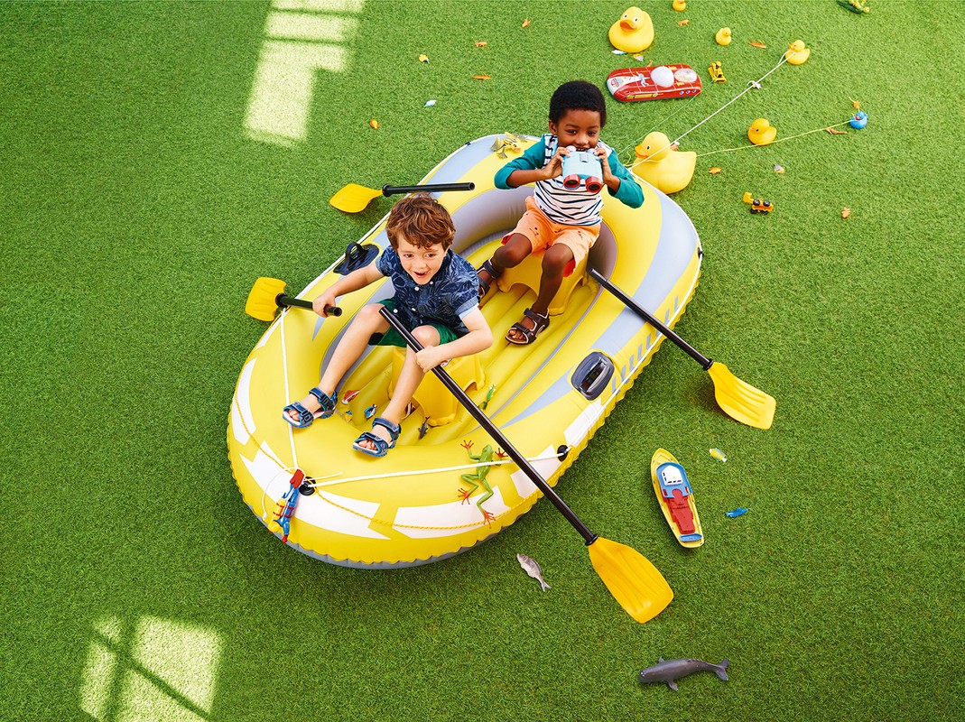 Two kids in a yellow inflatable raft on green grass surrounded by toy ducks, oars, toy shark, and small toy vehicles on a sunny day.