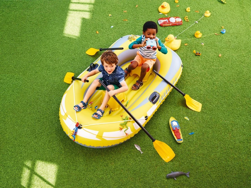 Two kids in a yellow inflatable raft on green grass surrounded by toy ducks, oars, toy shark, and small toy vehicles on a sunny day.