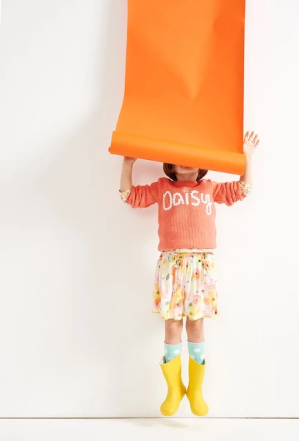 A young child holding an unrolled orange paper scroll over their head indoors, wearing a printed skirt, sweater, and yellow boots.