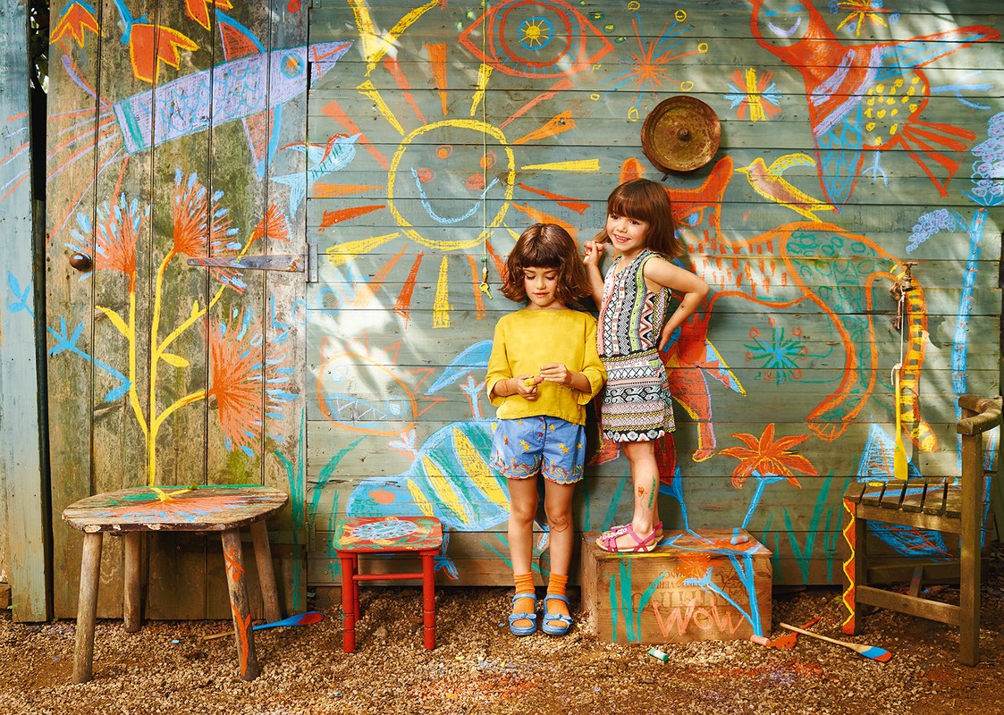 Two young girls pose in front of a colorful, rustic wooden wall adorned with chalk drawings of suns, flowers, and abstract shapes.