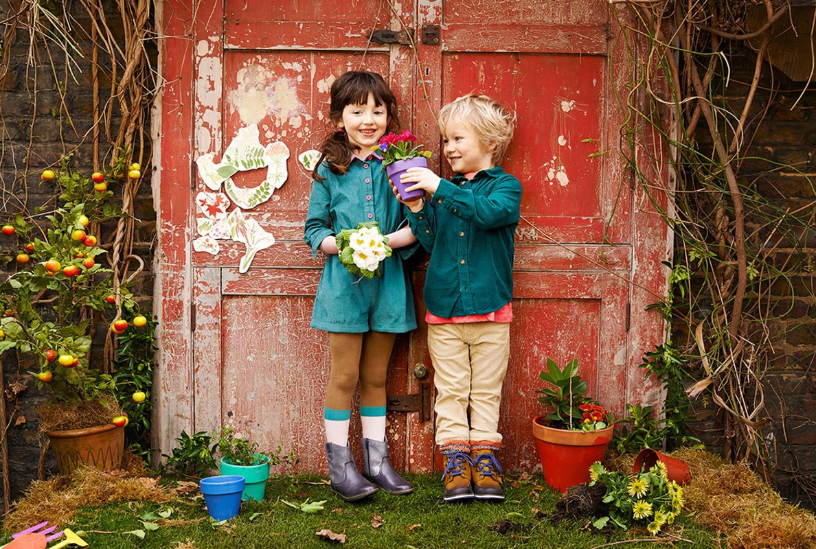 Two smiling children, a boy and a girl, stand in front of a weathered red door holding potted flowers, surrounded by plants and gardening props.