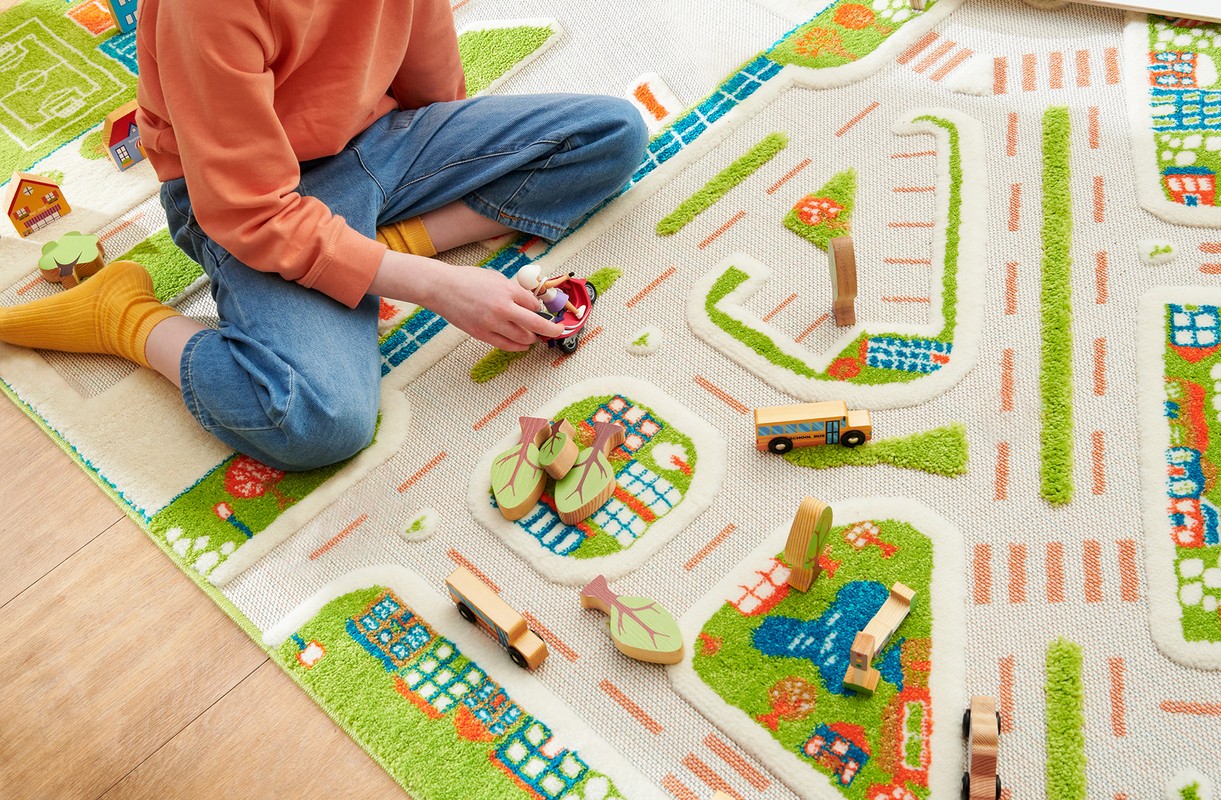 Child playing with toy cars on a colorful road-patterned rug indoors. The rug features roads, buildings, and trees in bright colors.