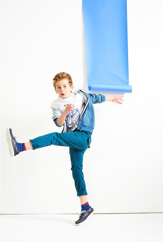 Full-length studio shot of a cheerful young boy in casual clothing kicking one leg and holding a blue paper roll against a white background.