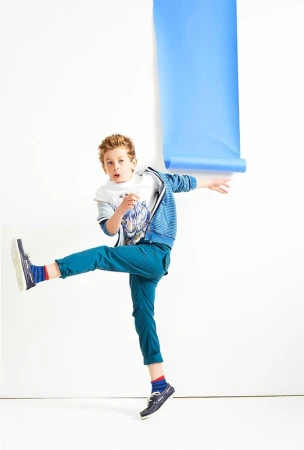 Full-length studio shot of a cheerful young boy in casual clothing kicking one leg and holding a blue paper roll against a white background.