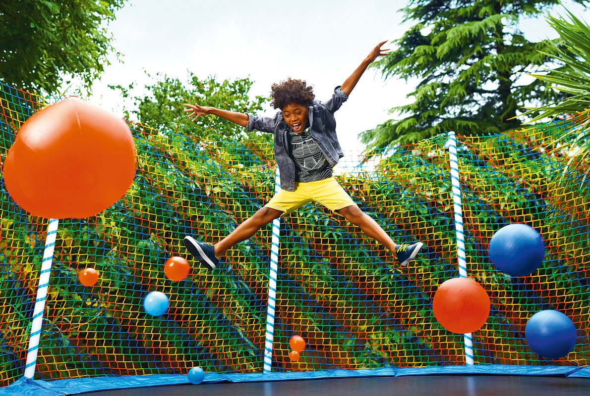 A young boy jumps high on a trampoline surrounded by floating orange and blue balls, set against a lush green background.