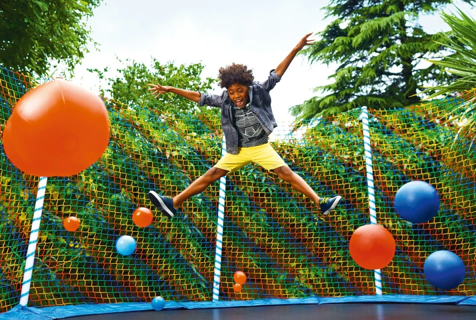 A young boy jumps high on a trampoline surrounded by floating orange and blue balls, set against a lush green background.