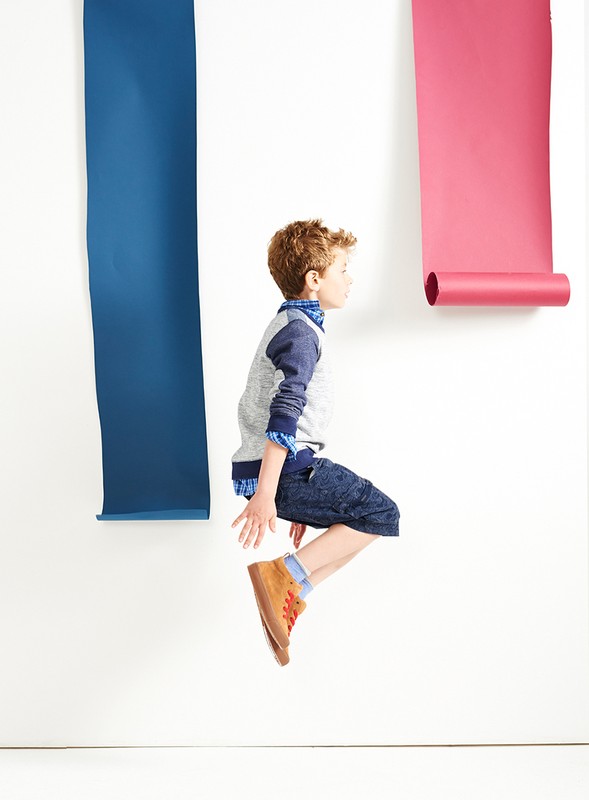 A young boy with curly red hair jumps in a photo studio against a white backdrop with blue and pink paper rolls attached.