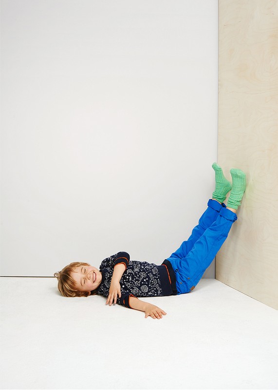 Child lying on floor, legs up against wall in blue pants and green socks, wearing a printed long-sleeve shirt, smiling at the camera.