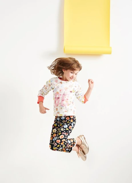 Young girl joyfully jumps in the air wearing patterned pants and a sweater, set against a clean white backdrop with a yellow paper roll.