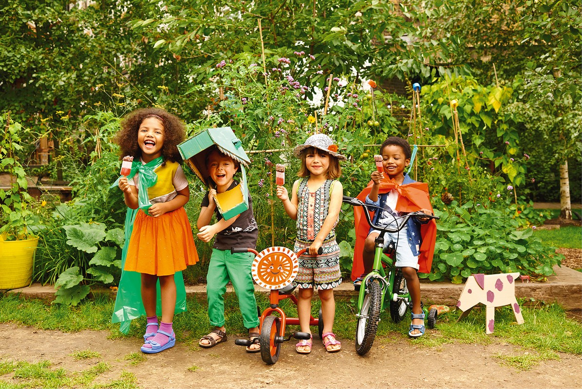 Four smiling children of varying ethnicities hold ice pops and wear homemade costumes with trees and garden greenery in the background.