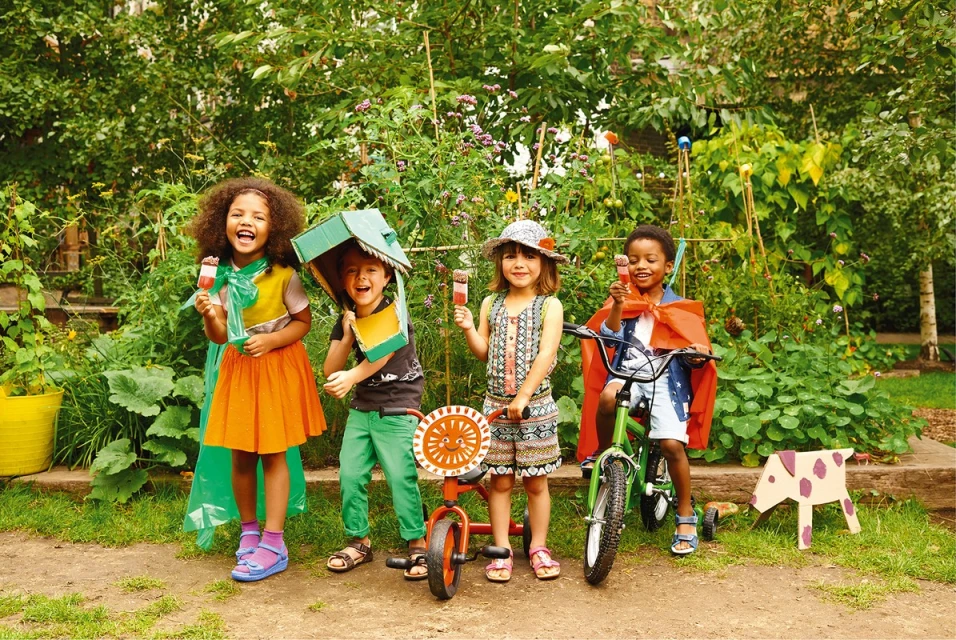 Four smiling children of varying ethnicities hold ice pops and wear homemade costumes with trees and garden greenery in the background.