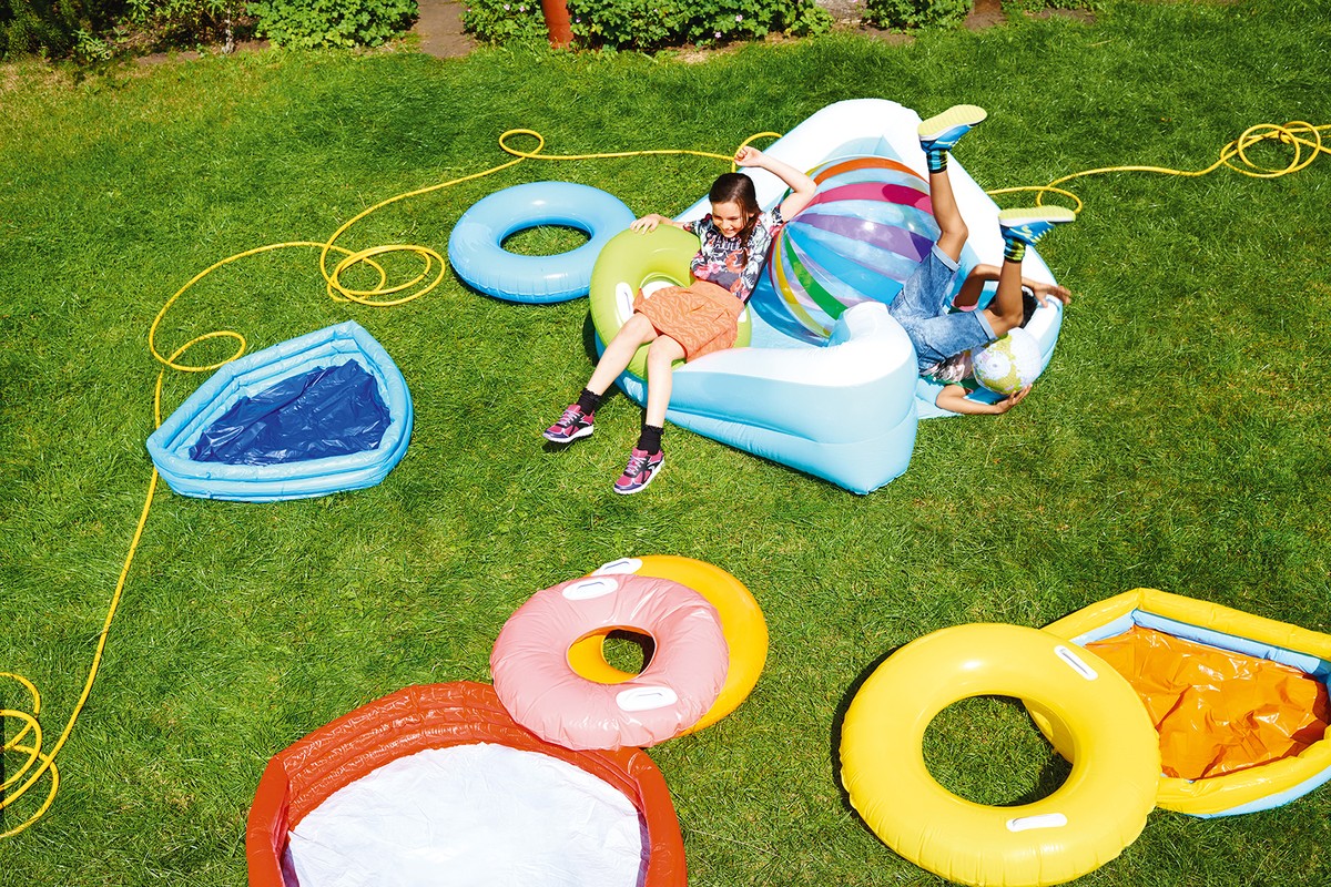 Two kids playing on an inflatable lounger outdoors surrounded by various colorful inflatable pool toys and a garden hose on a green lawn.