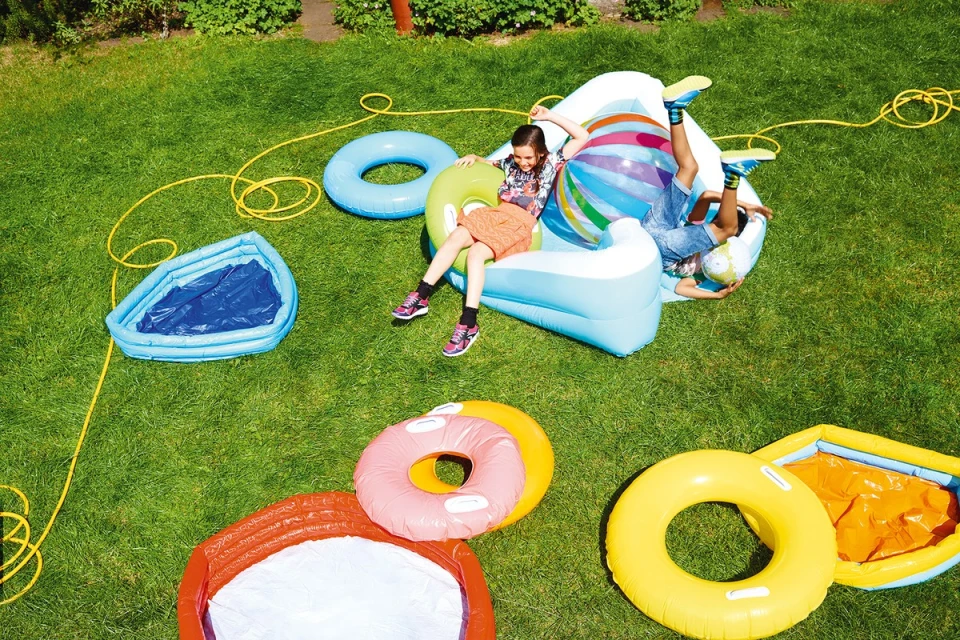 Two kids playing on an inflatable lounger outdoors surrounded by various colorful inflatable pool toys and a garden hose on a green lawn.