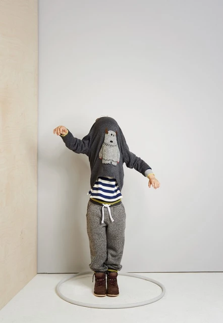 A child jumps inside a gray hula hoop in a gray and beige corner wearing brown boots, gray sweatpants, and a gray sweater over their head.