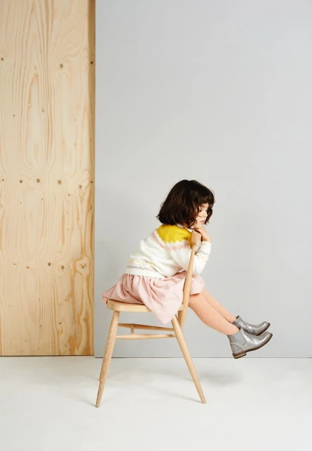 Young girl seated on a wooden chair, wearing a sweater, skirt, tights, and ankle boots in a minimalist studio setting.