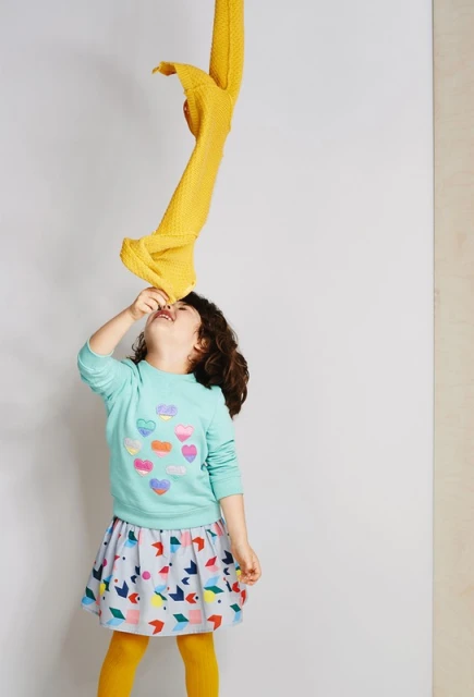 A young child with dark curly hair looks up at a yellow cloth suspended above, wearing a patterned skirt and colorful sweater indoors.