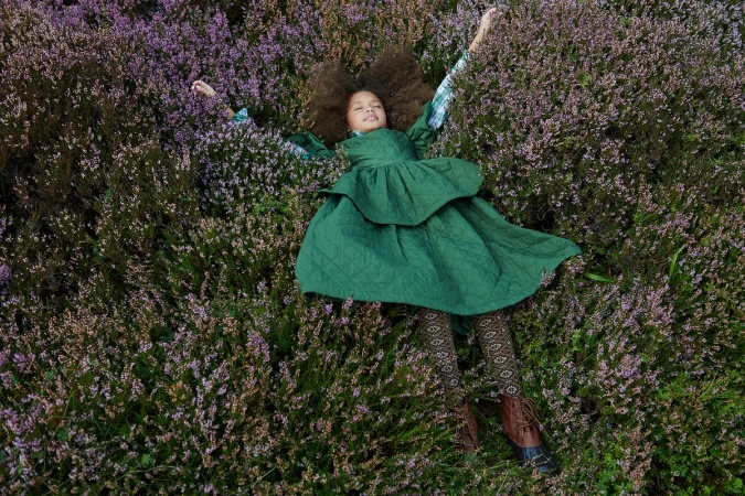A young girl with voluminous curly brown hair lies flat surrounded by purple heather flowers wearing a green quilted dress and patterned tights