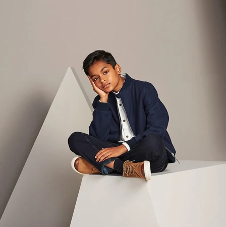 Young boy in a dark jacket, jeans and tan boots sits cross-legged on a white block in a photo studio with a pyramid in the background.