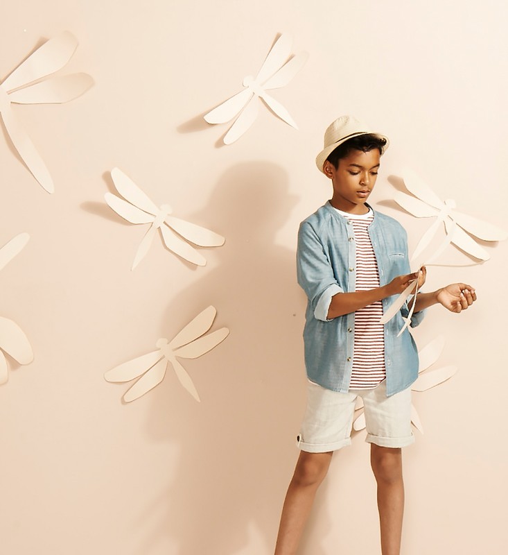 Young boy in a blue denim shirt and white shorts poses against a peach wall decorated with white dragonfly cutouts.