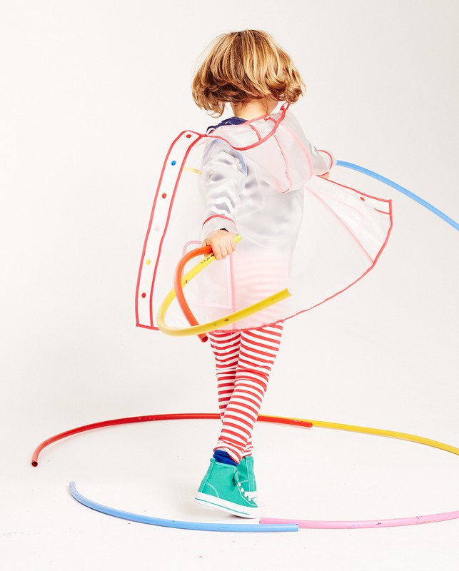 A child wearing a transparent raincoat with colored trim and striped leggings poses in a studio surrounded by colorful tubing.