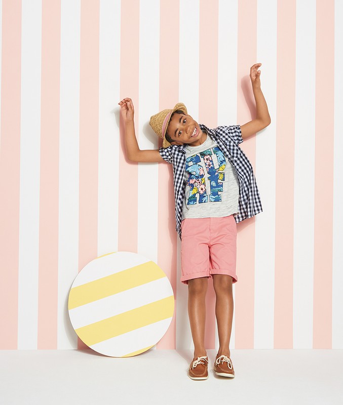 Young boy in preppy summer fashion poses against a striped wall with a circular prop, showcasing a playful and stylish look.