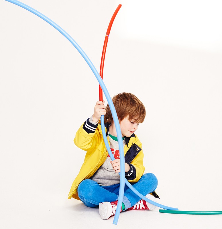 Young boy in a yellow jacket sits cross-legged, playfully interacting with colorful, flexible tubes in a brightly lit studio setting.