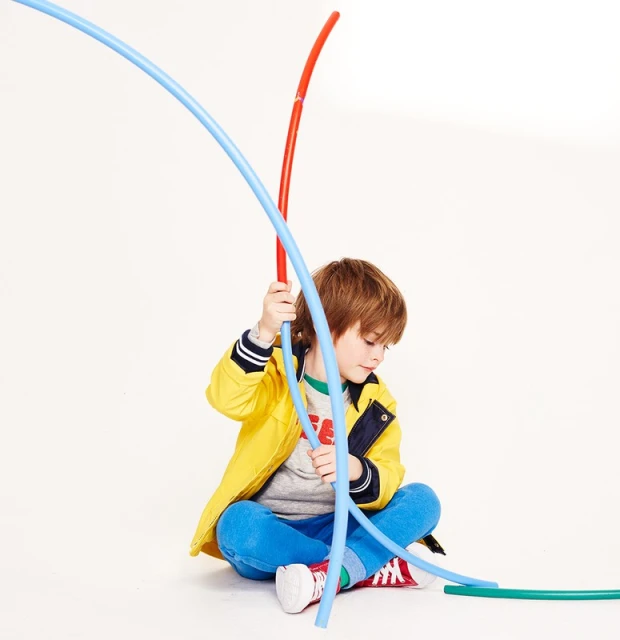 Young boy in a yellow jacket sits cross-legged, playfully interacting with colorful, flexible tubes in a brightly lit studio setting.