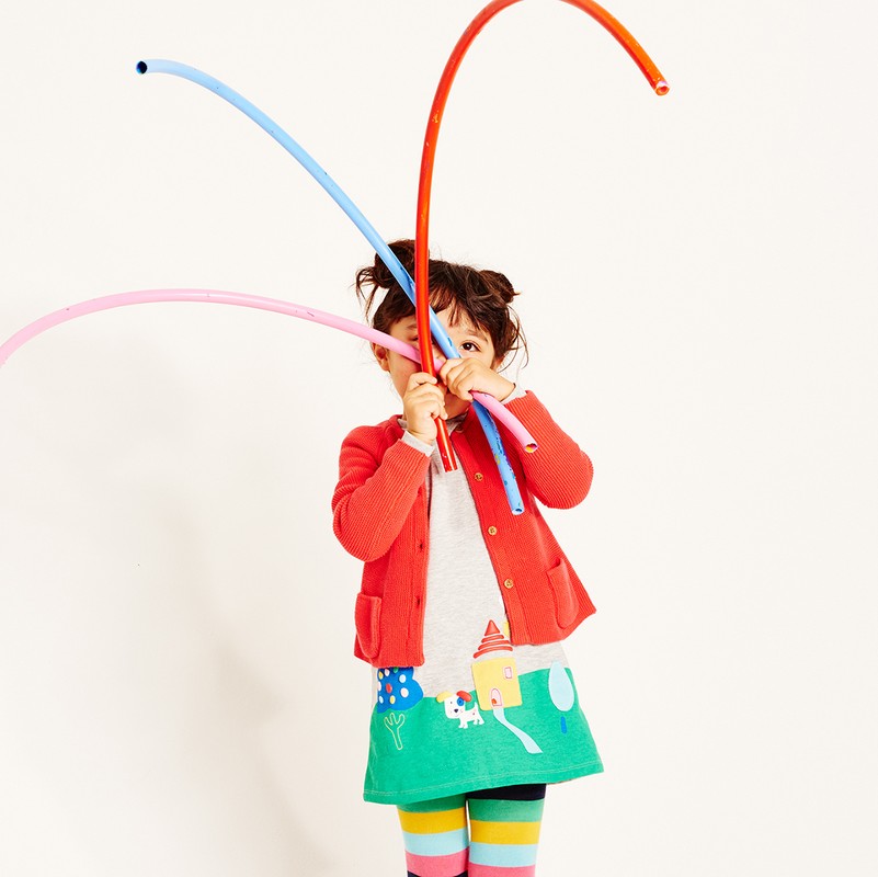 A young child plays with colorful flexible tubes against a white backdrop, wearing a red cardigan and a whimsical printed skirt, in a playful studio shot.