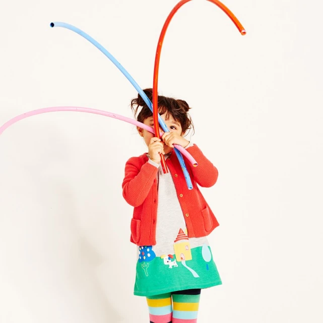 A young child plays with colorful flexible tubes against a white backdrop, wearing a red cardigan and a whimsical printed skirt, in a playful studio shot.
