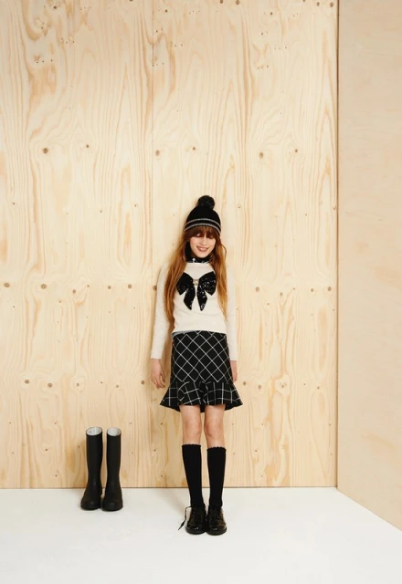 Full shot of a girl in winter fashion, posing in a long-sleeved top, checkered skirt, hat, and boots, against a plywood backdrop.