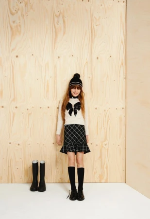 Full shot of a girl in winter fashion, posing in a long-sleeved top, checkered skirt, hat, and boots, against a plywood backdrop.