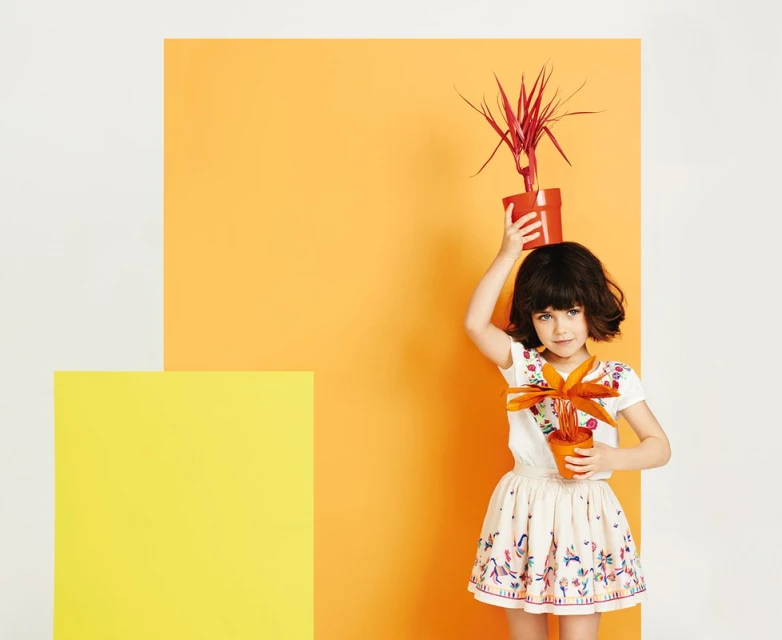 A young girl in a patterned skirt and white shirt poses indoors, holding a small potted plant with orange and red against color blocks.